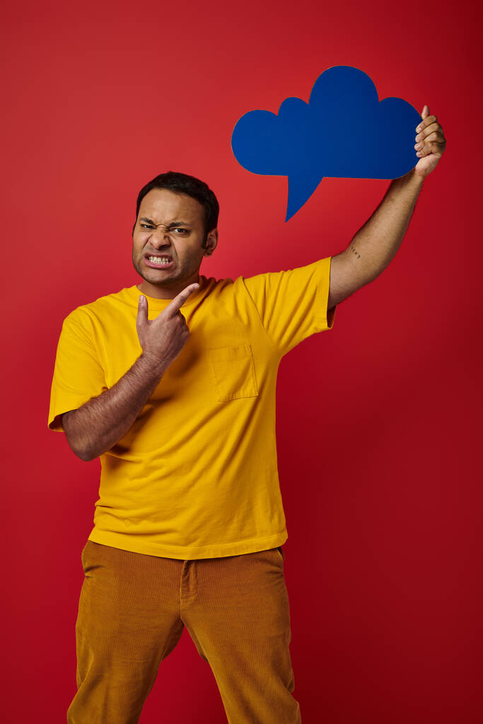 недовольный индиец в жёлтой футболке указывает пустым мыслей пузырь на красном фоне, гримаса лица - Фото, изображение