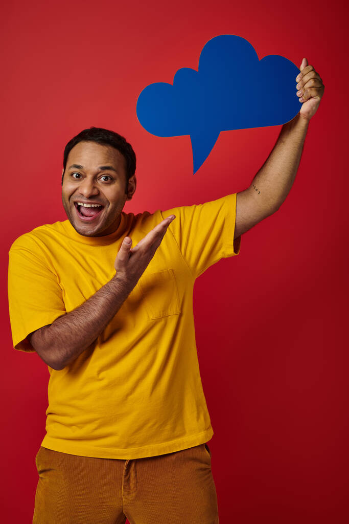 excited indian man in yellow t-shirt pointing blank thought bubble on red backdrop, happy face - Photo, Image