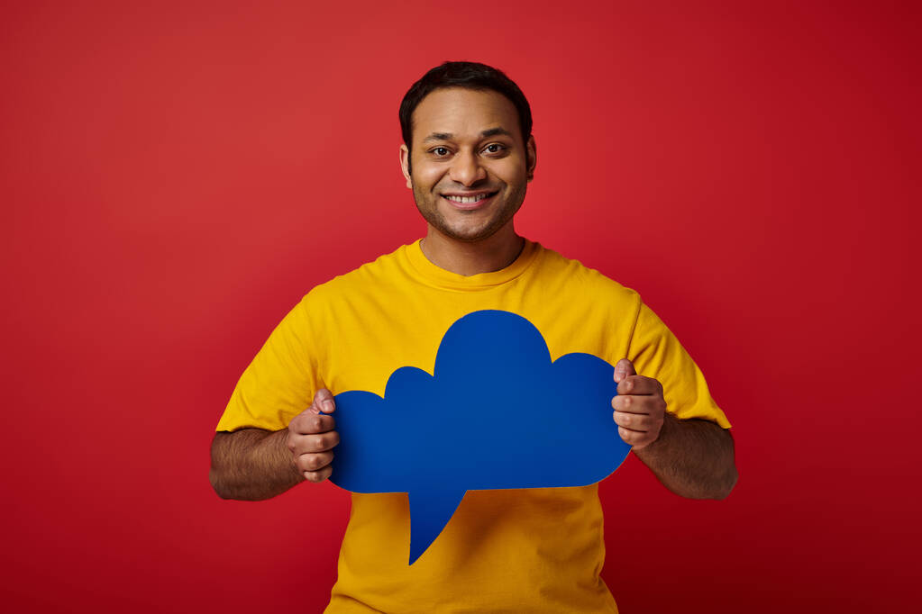 cheerful indian man in yellow t-shirt holding blue blank thought bubble on red backdrop, happy face - Photo, Image