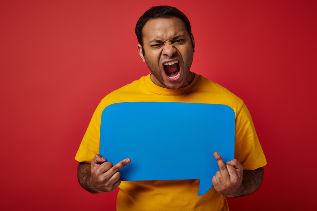 homem indiano provocante segurando bolha de fala em branco e mostrando os dedos do meio no fundo vermelho - Foto, Imagem