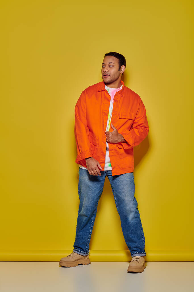 longueur totale de l'homme indien élégant en veste orange et jeans en denim debout sur fond jaune - Photo, image