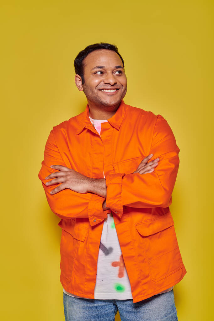 黄色い背景に折られた腕で立っているオレンジのジャケットの肯定的なインド人の男性の肖像画 - 写真・画像