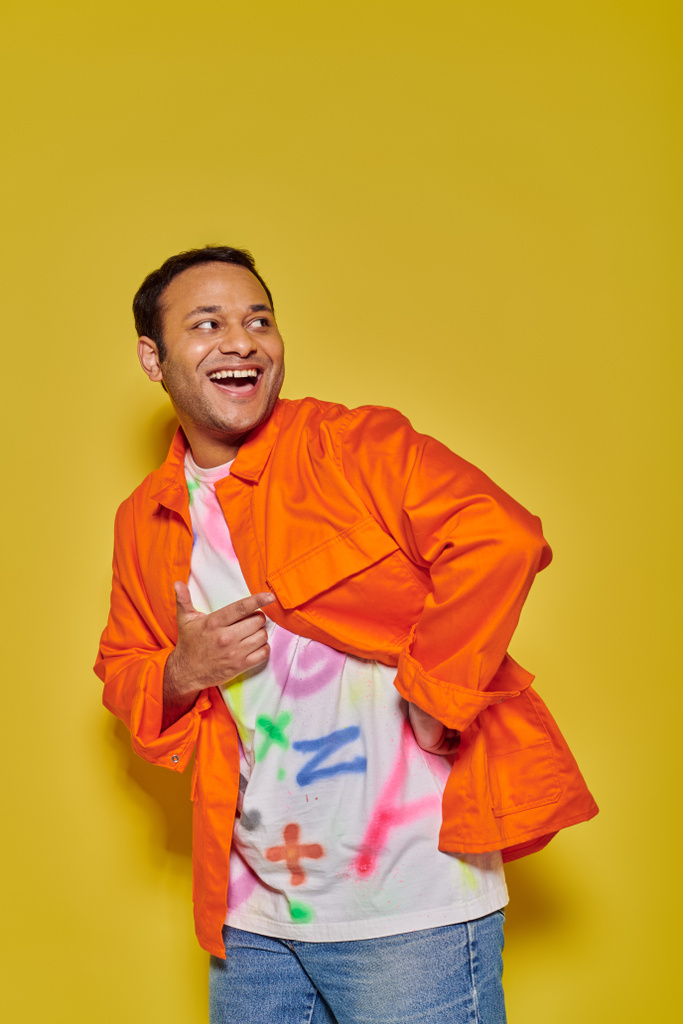 retrato de hombre indio excitado en chaqueta naranja y camiseta bricolaje mirando hacia otro lado en el fondo amarillo - Foto, Imagen