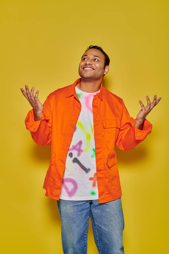 cheerful indian man in orange jacket and diy t-shirt gesturing on yellow backdrop, unbelievable - Photo, Image