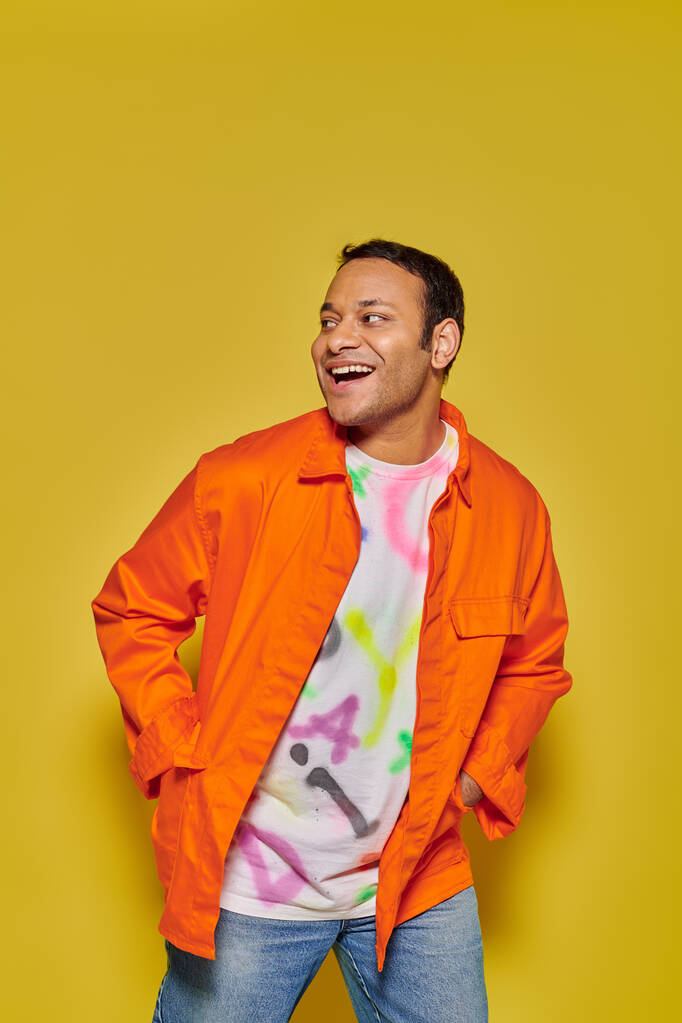 retrato de hombre indio alegre en chaqueta naranja posando con las manos en bolsillos sobre fondo amarillo - Foto, Imagen