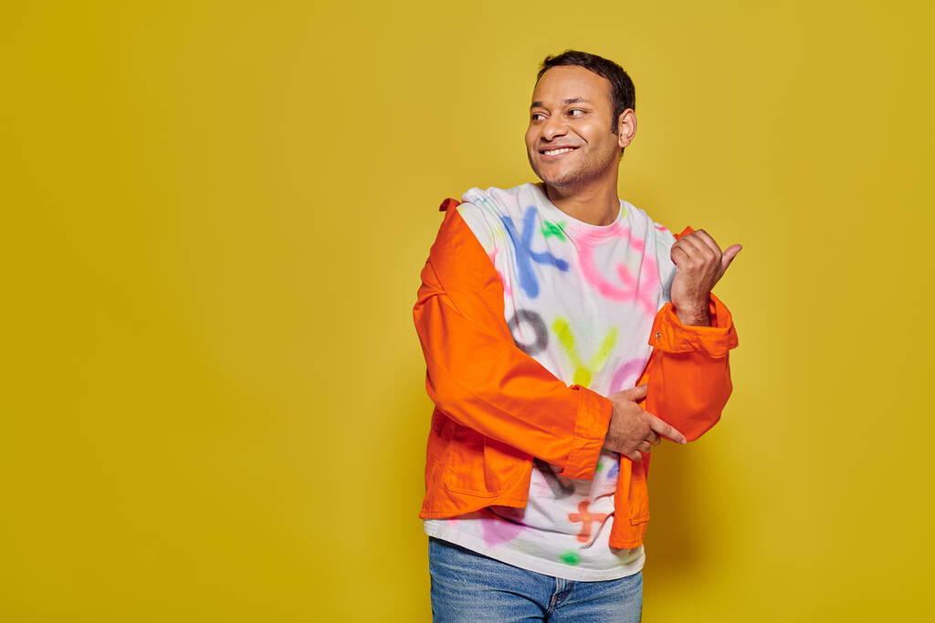 positive indian man in orange jacket and diy t-shirt pointing with thumb on yellow background - Photo, Image