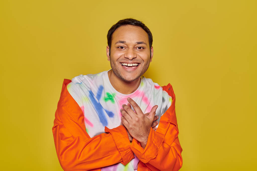 vrolijke indiaanse man in oranje jasje en diy t-shirt lachend en kijkend naar camera op gele achtergrond - Foto, afbeelding