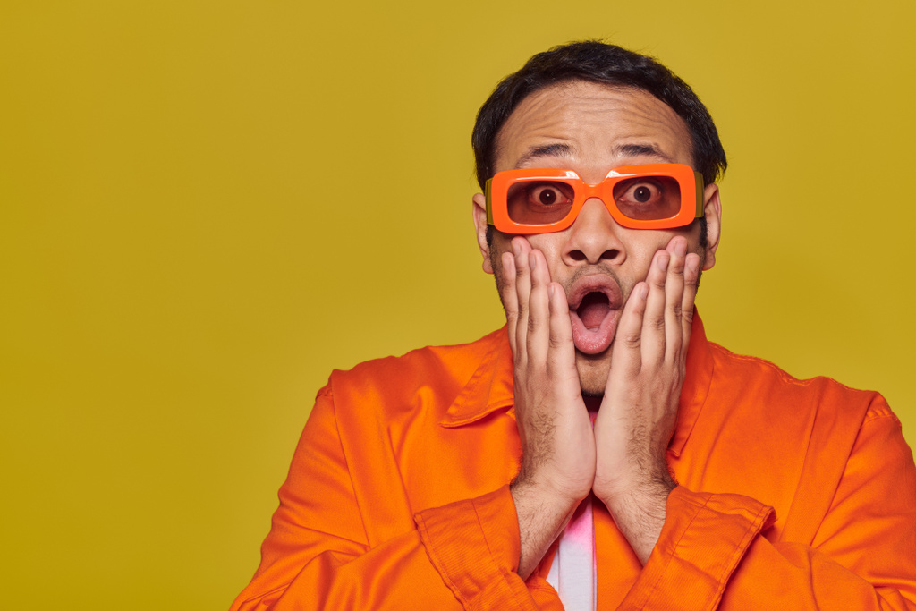 homme indien expressif et choqué en lunettes de soleil orange touchant le visage et disant wow sur fond jaune - Photo, image