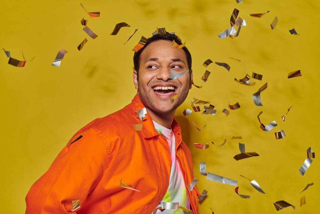 hombre indio alegre en chaqueta naranja brillante sonriendo cerca de confeti caída sobre fondo amarillo - Foto, Imagen