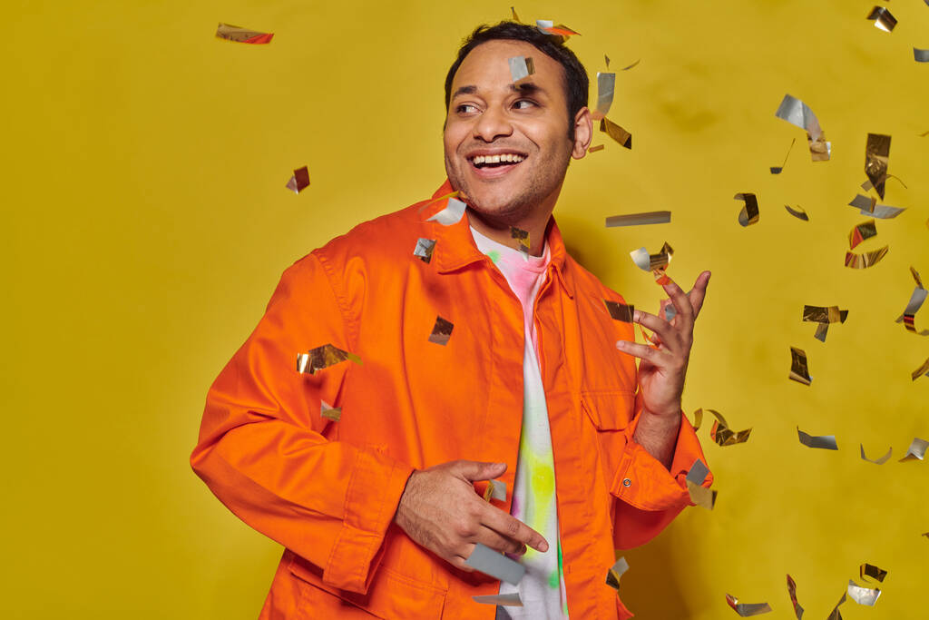 onnellinen intialainen mies kirkkaan oranssi takki hymyilee lähellä kuuluvat konfetti keltaisella taustalla, puolue - Valokuva, kuva