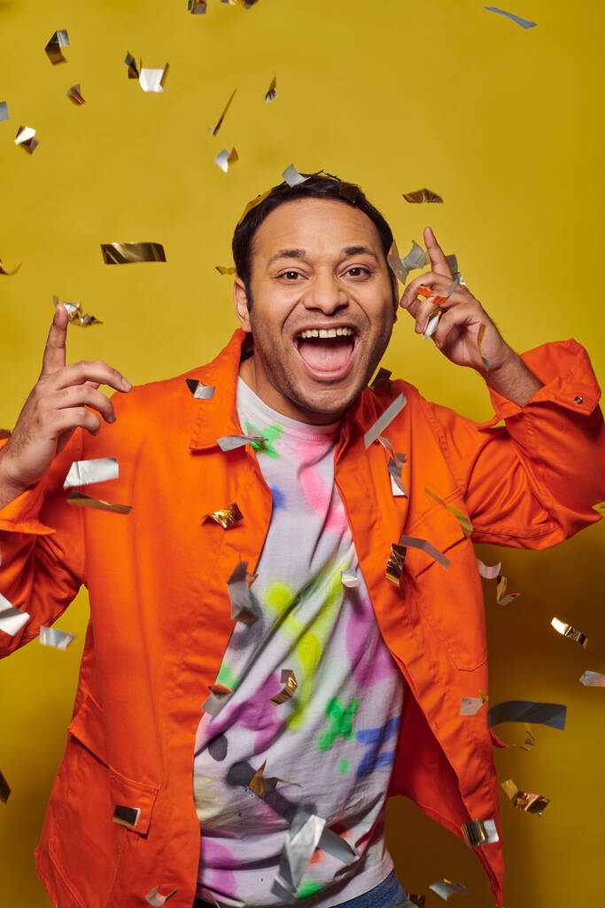 excité homme indien en veste orange souriant près de tomber confettis sur fond jaune, concept de fête - Photo, image