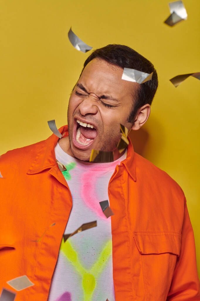 emotional indian man in orange jacket screaming near confetti on yellow backdrop, party - Photo, Image