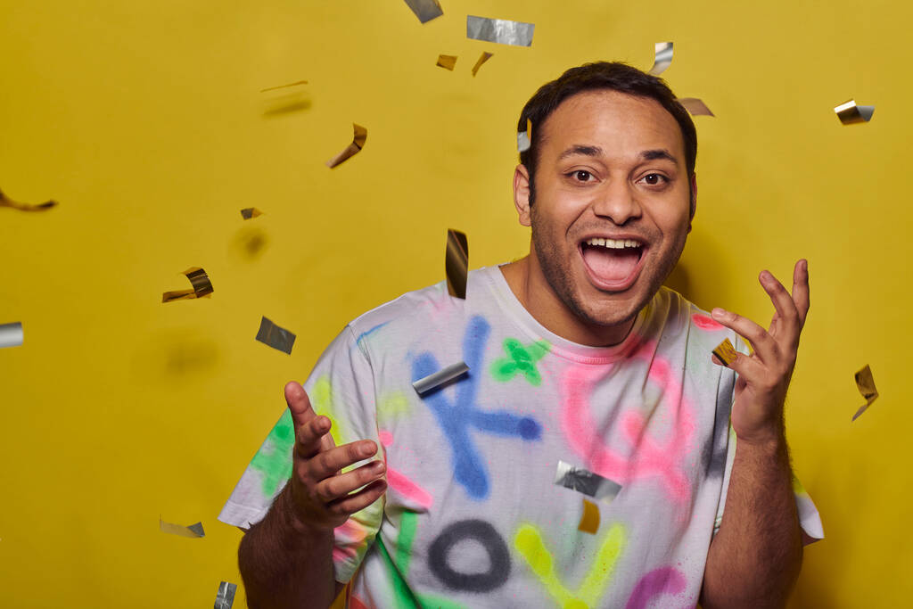 hombre indio feliz en camiseta de moda gesto cerca de caer confeti sobre fondo amarillo, concepto de fiesta - Foto, imagen