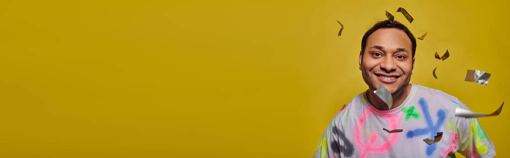 hombre indio positivo sonriendo cerca de caer confeti sobre fondo amarillo, concepto de fiesta, bandera - Foto, Imagen