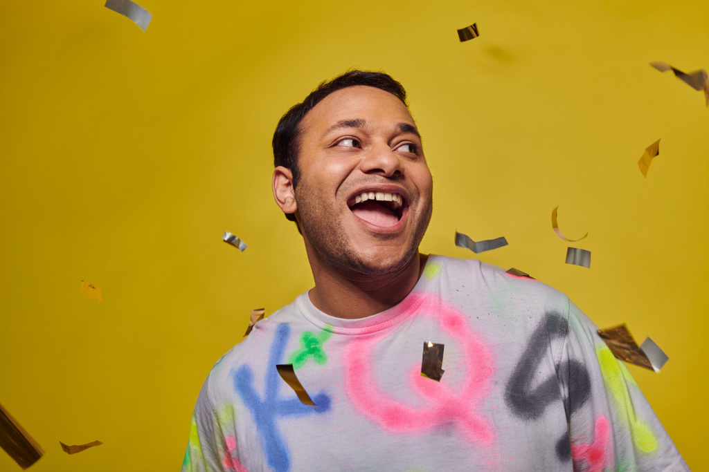 excited indian man smiling near falling confetti on yellow backdrop, party concept, happy face - Photo, Image