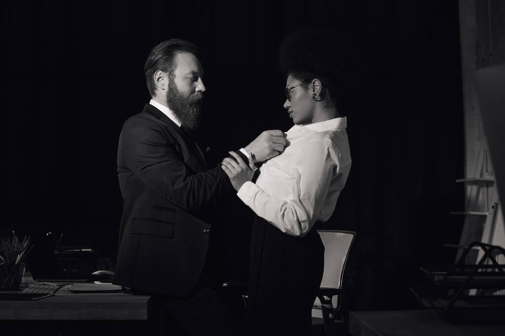 Schwarz-Weiß-Foto eines Geschäftsmannes beim Ausziehen einer afrikanisch-amerikanischen Frau im Nachtbüro, Seitenansicht - Foto, Bild