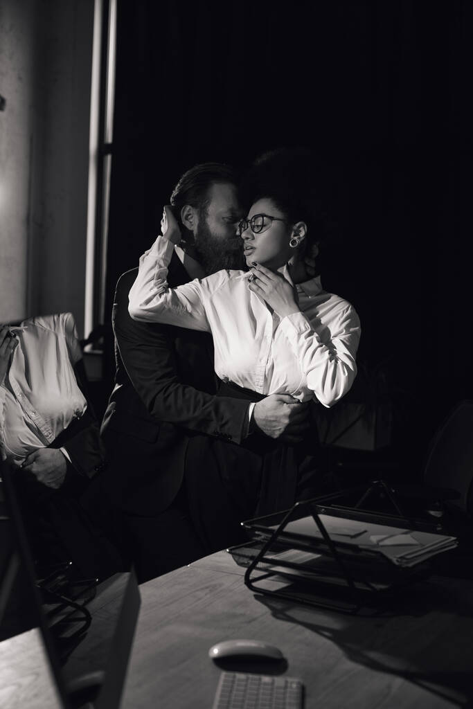 interrassische Paar in formeller Kleidung umarmen sich im Büro in der Nacht, Liebesaffäre, Schwarz-Weiß-Foto - Foto, Bild