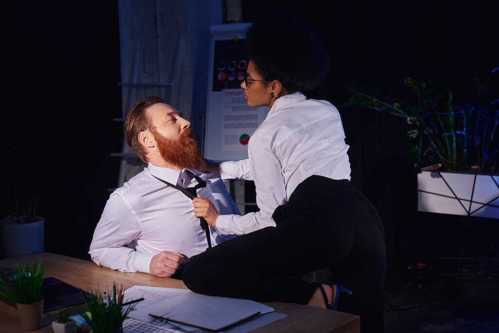 young african american businesswoman pulling tie of bearded businessman at work desk, office romance - Photo, Image