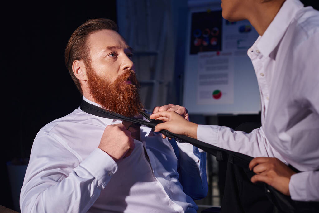 изумленный бородатый бизнесмен рядом с африканской американкой дергает за галстук, соблазняет в темном кабинете - Фото, изображение