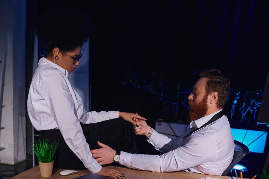 vousatý obchodník držící se za ruku africké Američanky sedící na stole, romantika v noční kanceláři - Fotografie, Obrázek
