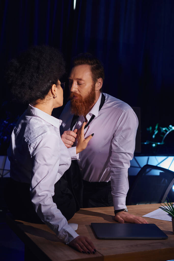 charismatic bearded businessman undressing near young african american woman in office at night - Photo, Image