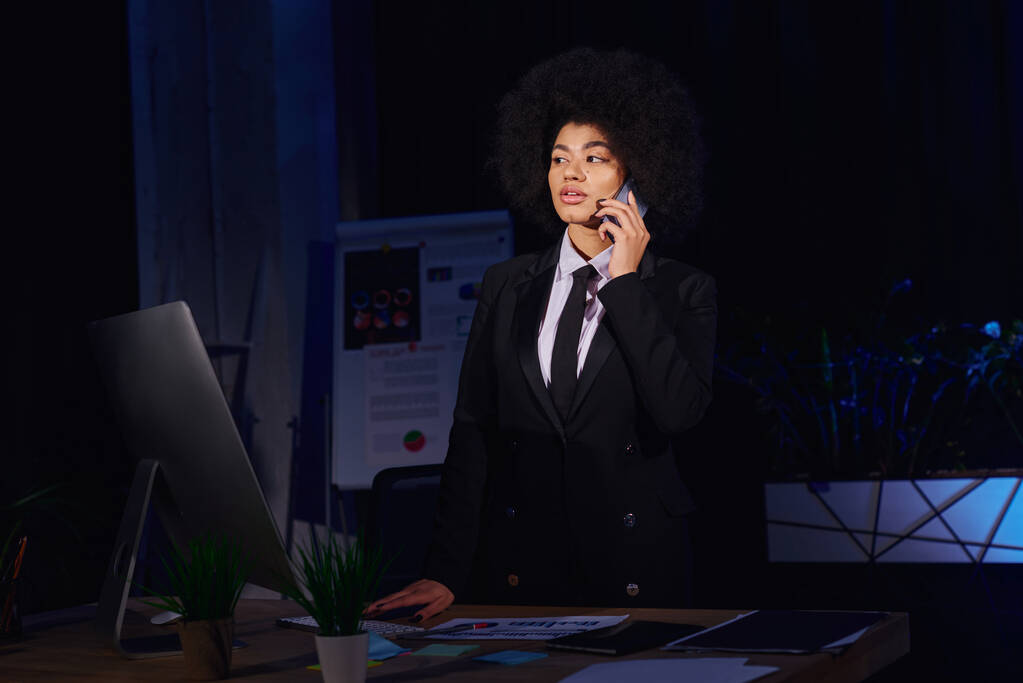 afro-américaine femme d'affaires parlant sur smartphone près de l'écran d'ordinateur dans le bureau la nuit - Photo, image