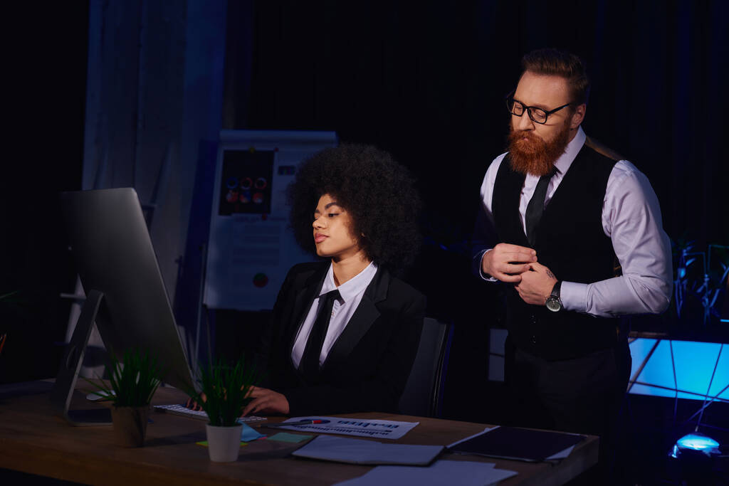бородатый бизнесмен смотрит на довольно африканский американский секретарь печатает на компьютере в ночном офисе - Фото, изображение