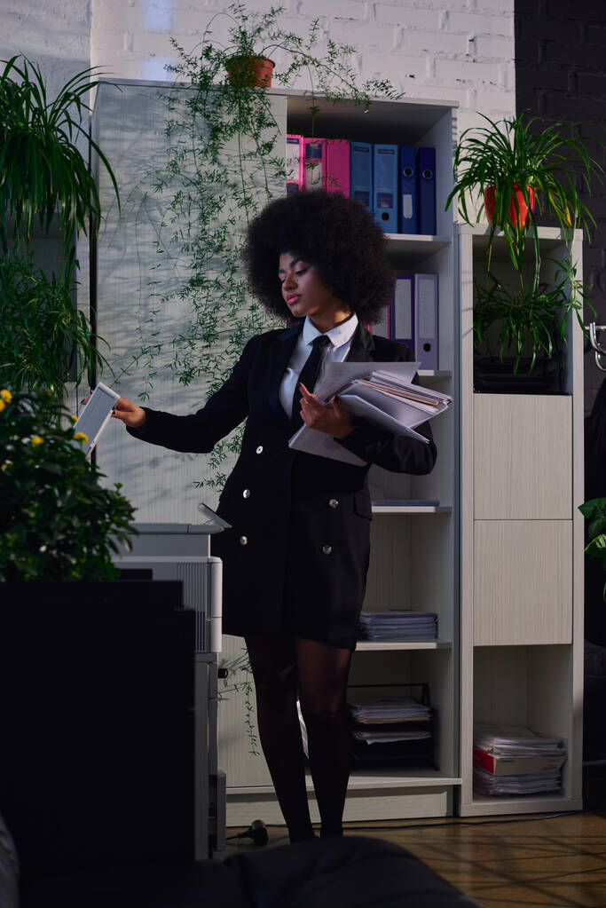 Afrikaans amerikaanse vrouw in elegante formele dragen kopiëren documenten tijdens het werken 's nachts in het kantoor - Foto, afbeelding
