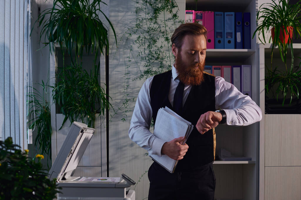 γενειοφόρος επιχειρηματίας με χαρτιά που κοιτάζει ρολόι κοντά στο φωτοτυπικό ενώ εργάζεται μέχρι αργά στο γραφείο - Φωτογραφία, εικόνα