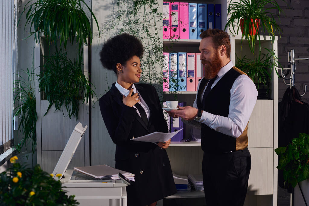 bearded businessman holding coffee cup and flirting with african american secretary in office - Photo, Image