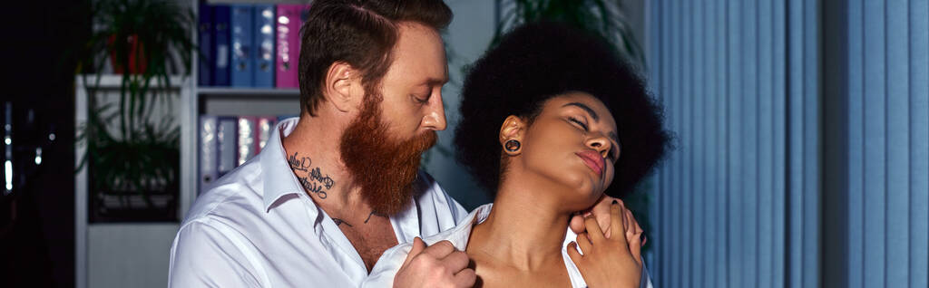 hombre de negocios barbudo tatuado seduciendo a secretaria afroamericana con los ojos cerrados en la oficina, pancarta - Foto, imagen