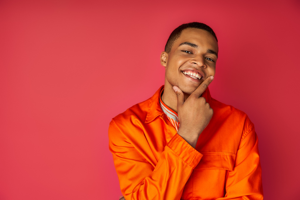 hombre afroamericano feliz en camisa naranja tocando la cara y mirando hacia otro lado sobre fondo rojo - Foto, imagen