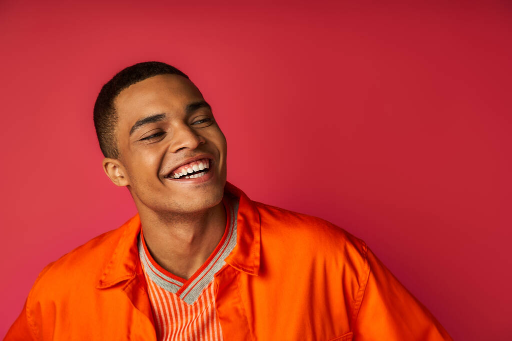 portrait of stylish and positive african american guy in orange shirt looking away on red background - Photo, Image