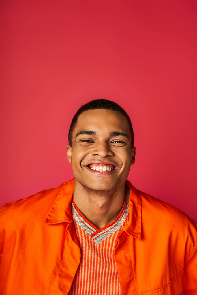 optimistische Afrikaans-Amerikaanse man lacht naar camera op rode achtergrond, oranje shirt, portret - Foto, afbeelding