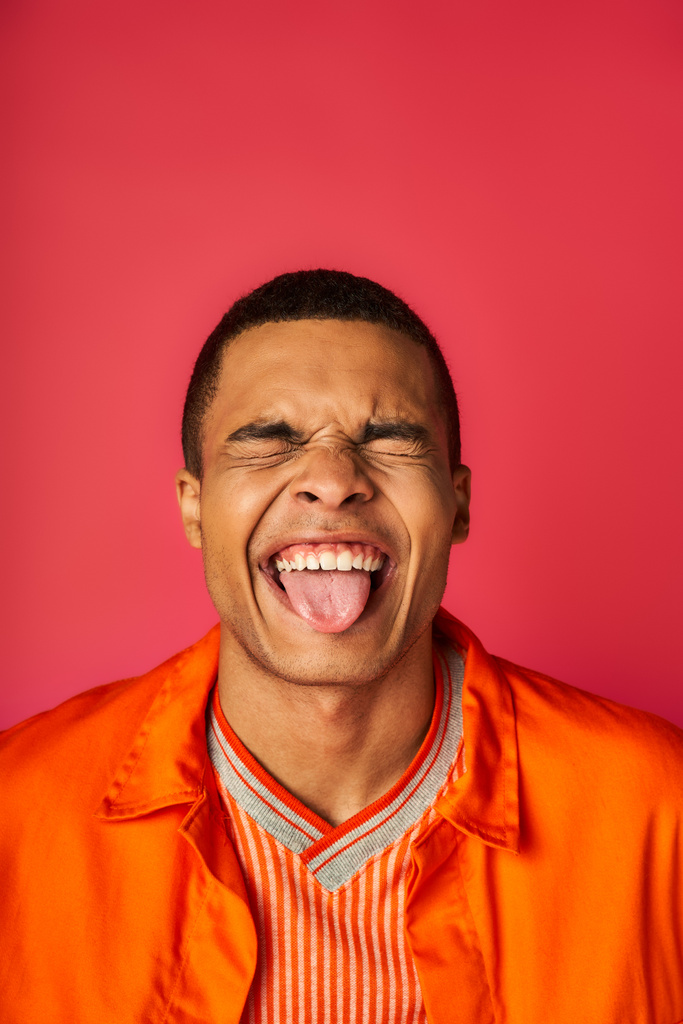 divertente afroamericano con gli occhi chiusi che sporgono lingua su sfondo rosso, camicia arancione, elegante - Foto, immagini