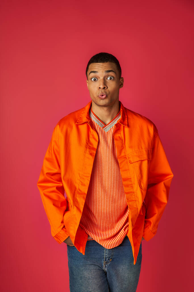 ontmoedigd Afrikaans-Amerikaanse man pruilend lippen en kijkend naar camera op rood, oranje shirt, stijlvol - Foto, afbeelding