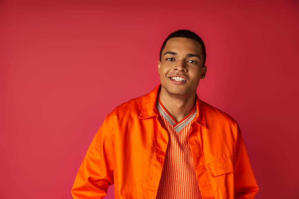 knap Afrikaans amerikaans man in trendy oranje shirt, met stralende glimlach kijken naar camera op rood - Foto, afbeelding