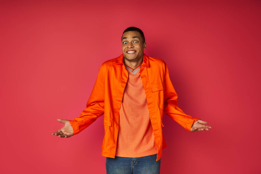 uomo afroamericano scoraggiato in camicia arancione mostrando gesto scrollata di spalle su sfondo rosso - Foto, immagini