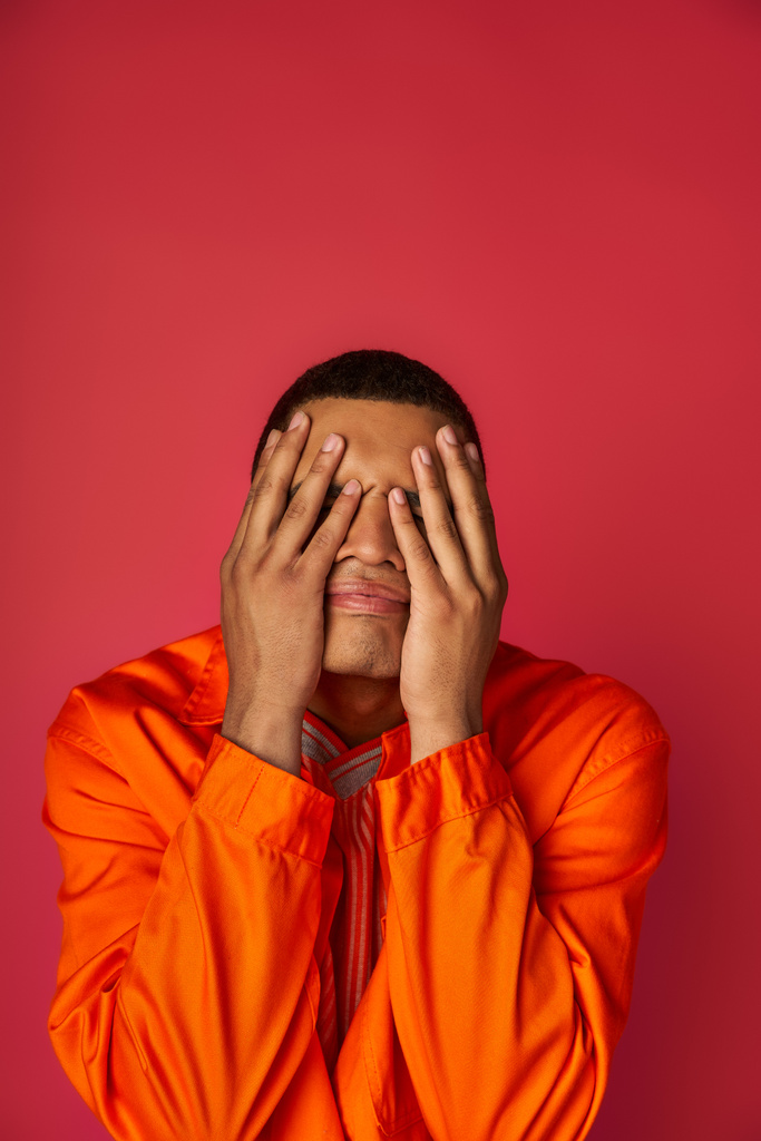 homme afro-américain troublé en chemise orange visage obscurcissant avec les mains sur fond rouge - Photo, image