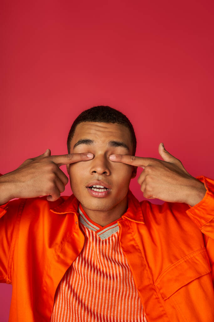 homem americano africano positivo em camisa laranja obscurecendo os olhos com os dedos no fundo vermelho - Foto, Imagem