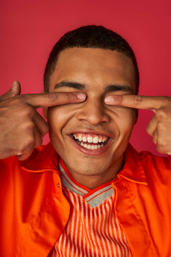 hombre afroamericano divertido oscurecer los ojos con los dedos, camisa naranja, fondo rojo - Foto, Imagen