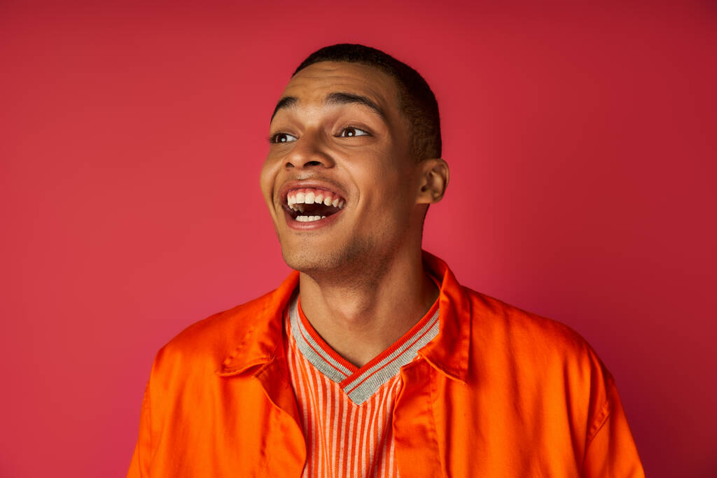 κατάπληκτος Αφροαμερικάνος με πορτοκαλί πουκάμισο κοιτάζει αλλού και γελάει με κόκκινο φόντο - Φωτογραφία, εικόνα