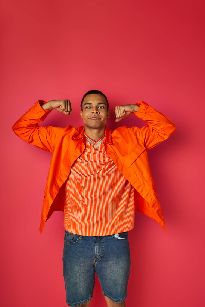 hombre afroamericano confiado en camisa naranja demostrando fuerza sobre fondo rojo - Foto, imagen