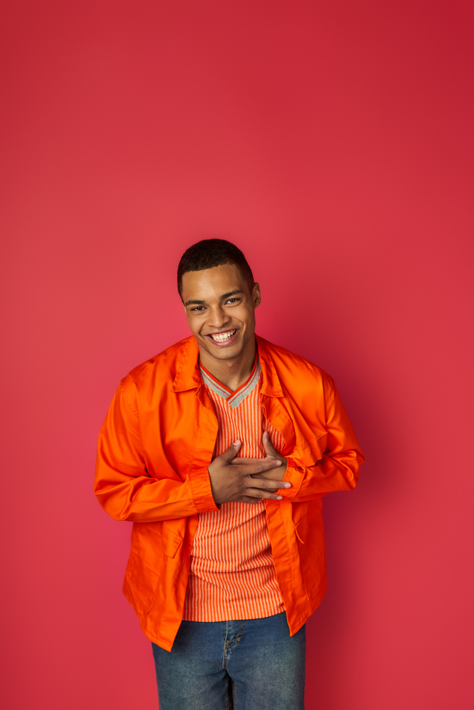 рад африканский американец трогает грудь, улыбаясь в камеру на красном, благодарный жест, оранжевая рубашка - Фото, изображение