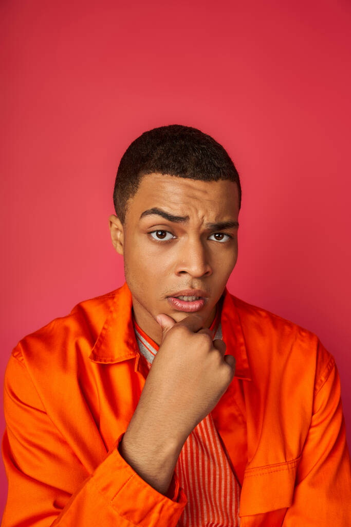 homme afro-américain réfléchi et inquiet en chemise orange touchant la poitrine, regardant la caméra sur le rouge - Photo, image
