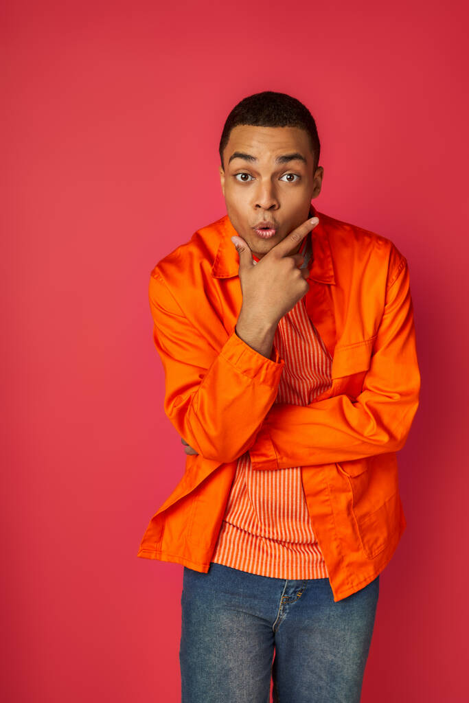 взволнованный африканский американец в оранжевой рубашке, с рукой возле лица, смотрящей в камеру на красном - Фото, изображение