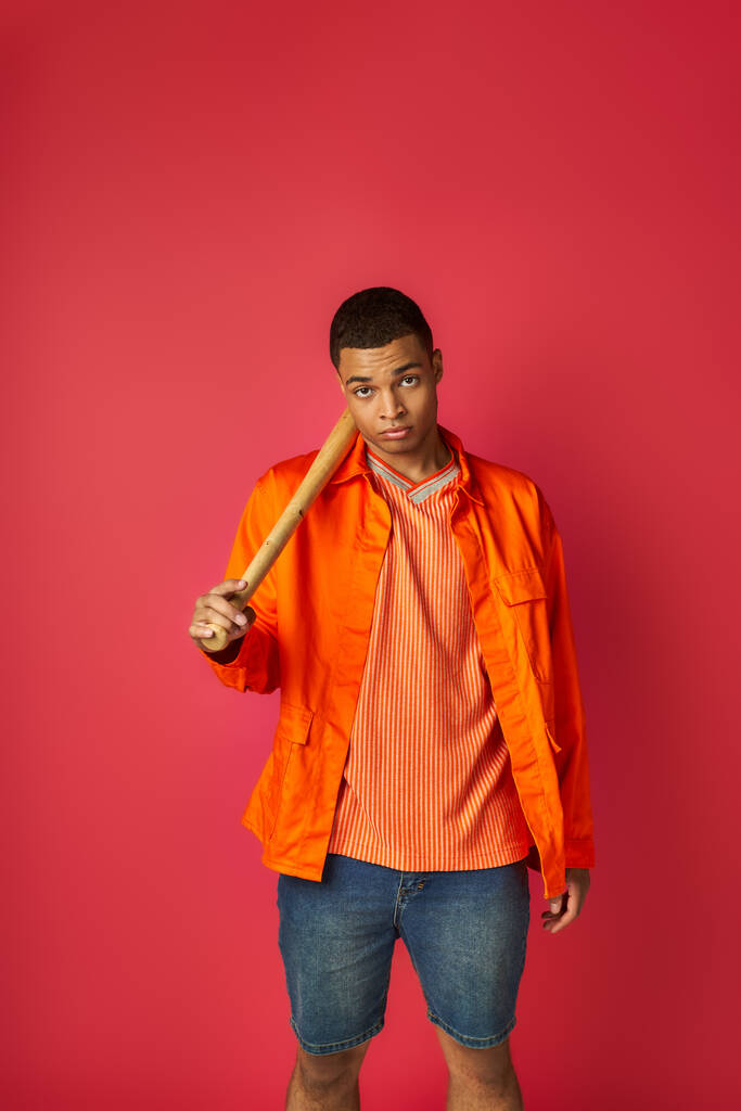 confiado hombre afroamericano en camisa naranja sosteniendo bate de béisbol y mirando a la cámara en rojo - Foto, Imagen