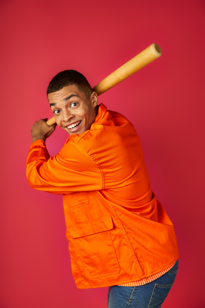 energizado afroamericano en camisa naranja jugando béisbol sobre fondo rojo - Foto, Imagen