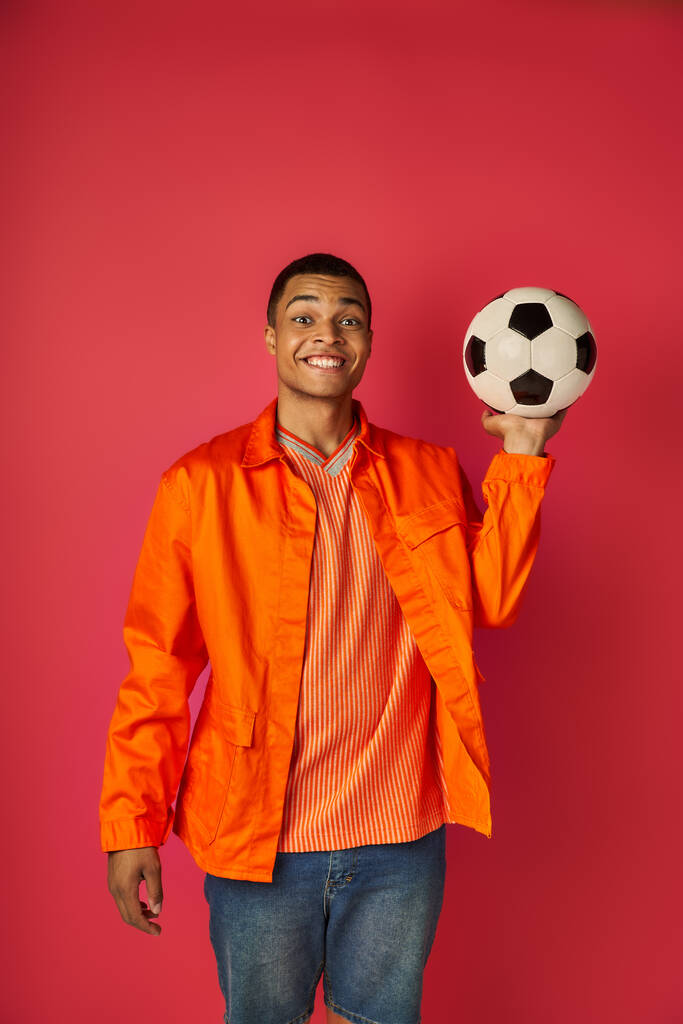 cheerful african american man in orange shirt holding soccer ball and smiling at camera on red - Photo, Image
