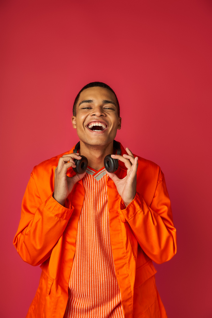 hombre afroamericano con estilo en camisa naranja, con auriculares inalámbricos riendo en rojo - Foto, Imagen